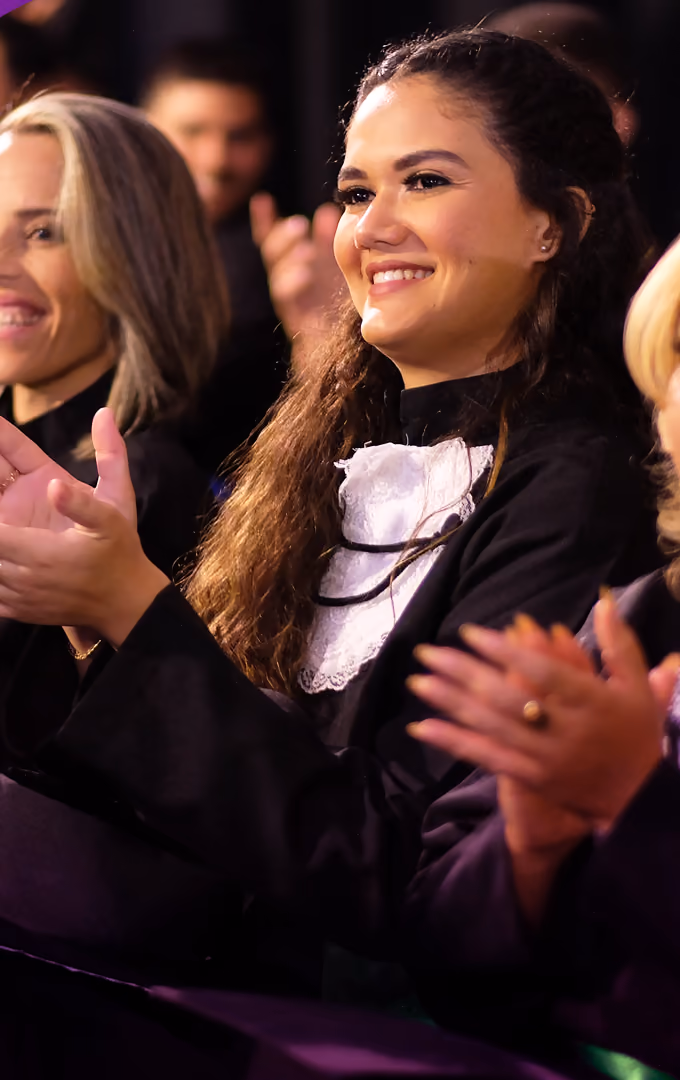 Formanda de beca aplaude sentada na plateia durante a cerimônia, ao lado de outras convidadas.