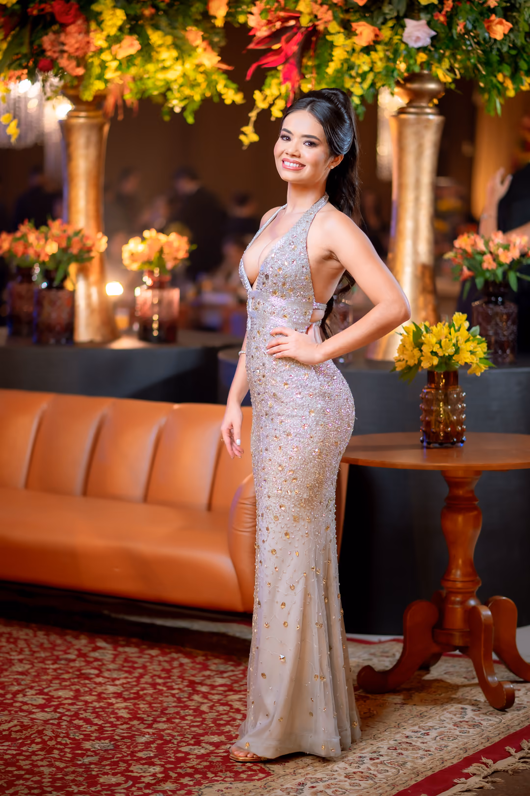Formanda em vestido longo prateado no salão do baile de gala, sob a decoração floral suspensa.