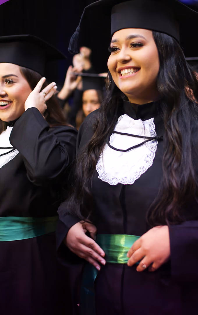 Duas formandas de beca com faixa verde ajustam o capelo e sorriem antes da entrada da cerimônia.
