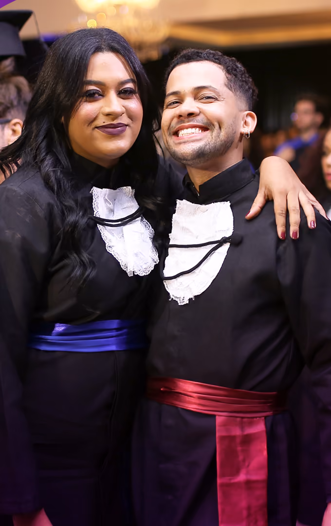Dois formandos de becas com faixas de cores diferentes, de cursos distintos, abraçados e sorrindo durante a colação unificada.