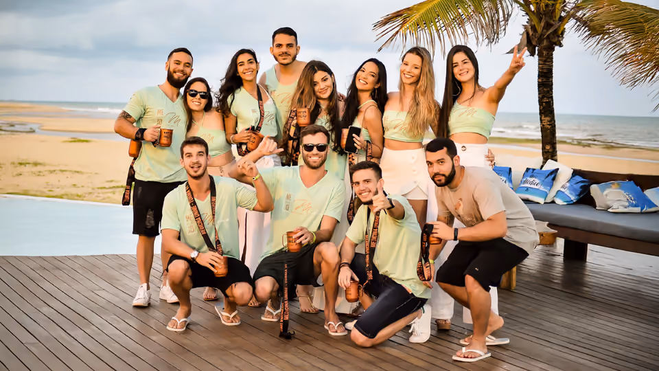 Turma reunida para foto em um pré-evento na praia, de camiseta personalizada, com coqueiro e mar ao fundo.